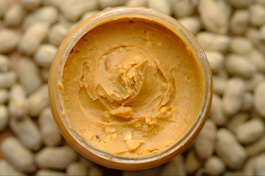 A top view of a jar of peanut butter sitting amidst whole peanuts in the shell.